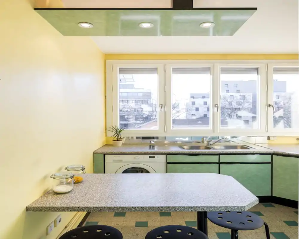 Bright compact kitchen with a corner sink, green cabinets and a small breakfast counter facing large windows that provide city views and plenty of natural light.