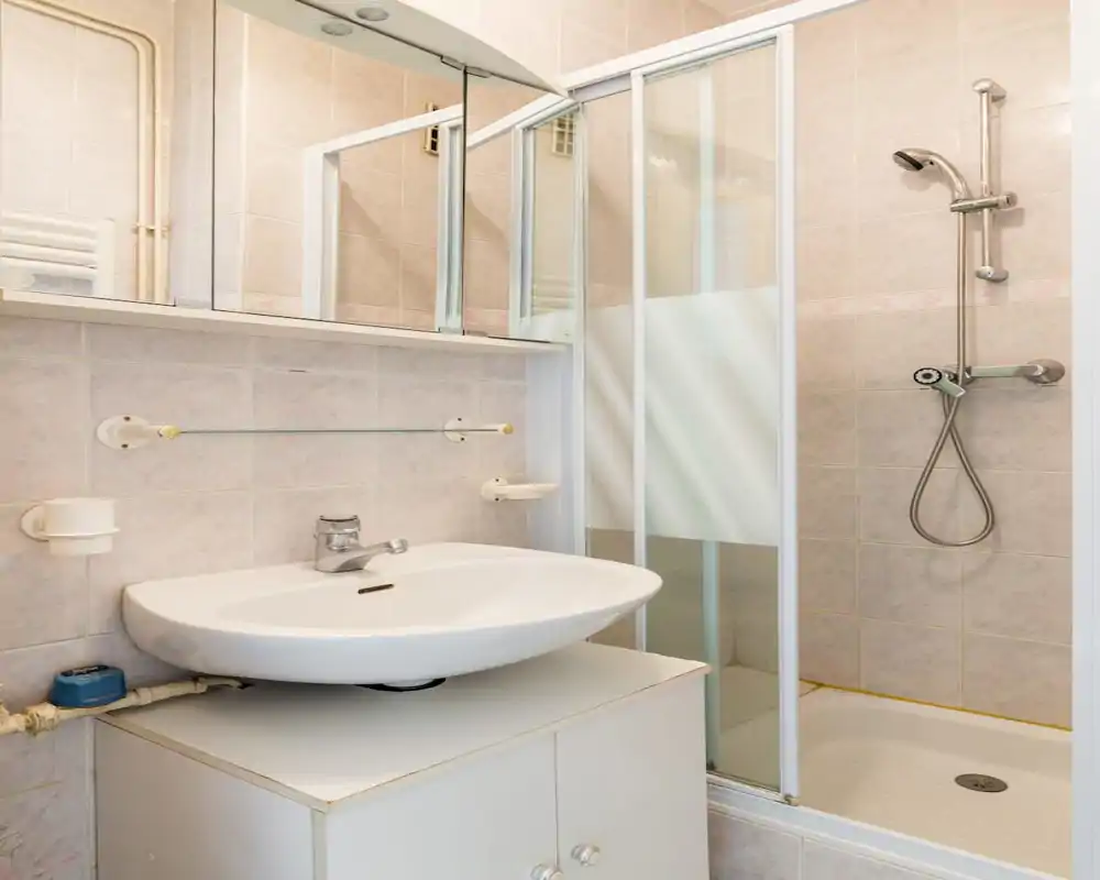 Compact, well-lit bathroom with a wall-mounted sink, mirrored cabinet and a corner shower with sliding glass doors — tidy and functional for a small rental.