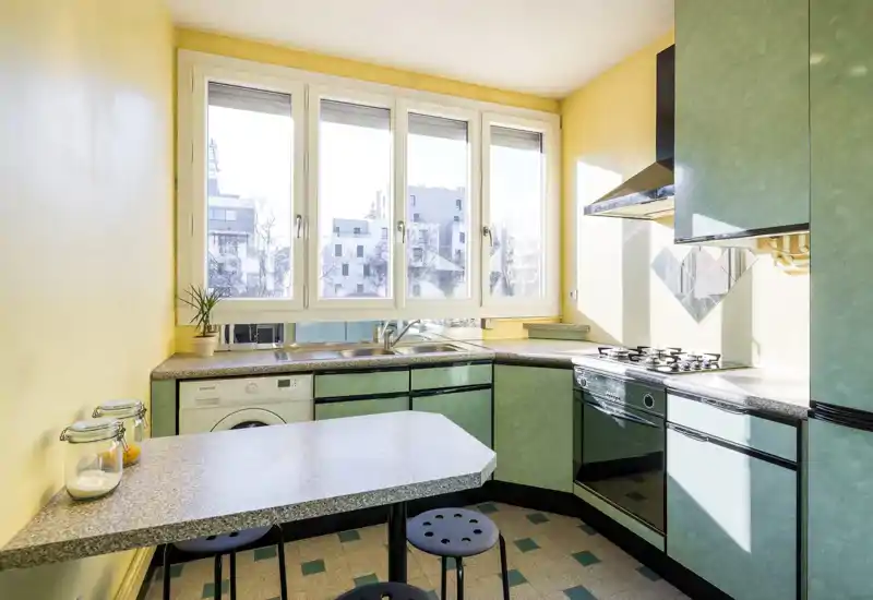 Bright, compact kitchen with green cabinets, a built-in oven and four-burner stove, a double sink under large windows and a small breakfast counter — well lit and neatly presented.
