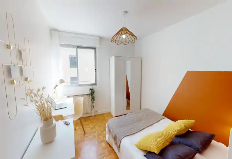 Bright, modern bedroom with a comfortable double bed, warm orange headboard accent and cozy textiles. Natural light fills the room from a large window, and a small desk creates a functional workspace.