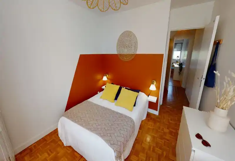 Sunny, well-staged bedroom with a bold terracotta accent wall, comfortable double bed dressed with white linens, yellow cushions and a patterned throw. Warm bedside lamps and natural light make the room inviting and cozy — ideal for rental photos.