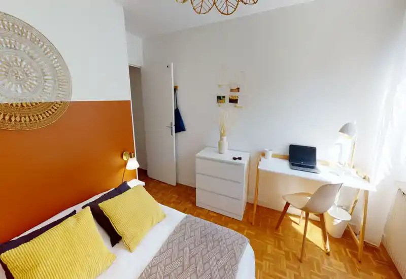 Bright, stylish bedroom with a large bed dressed in yellow cushions and a woven throw. A desk by the window provides a sunny workspace and a minimalist chest of drawers adds storage.