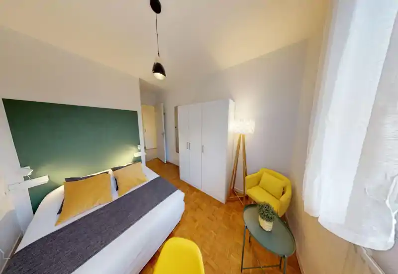 Bright, well-staged bedroom with a large bed framed by a green headboard, warm accents and a cozy reading corner with a yellow armchair and floor lamp.