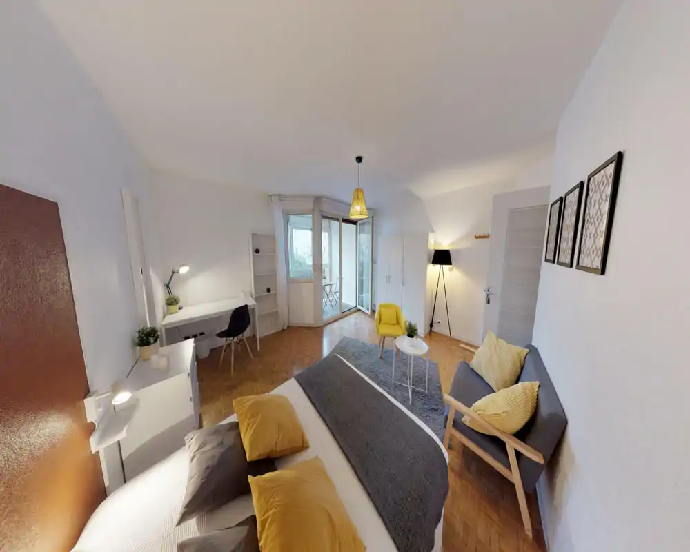 Bright, modern bedroom with a large dressed bed in the foreground, cozy textiles and pillows, a small seating area and desk, and sliding doors leading to a balcony.