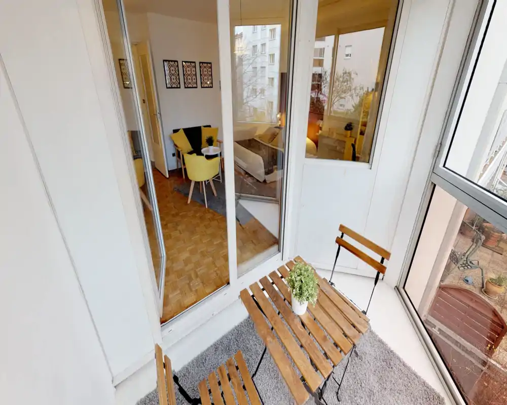 Sunny enclosed balcony with a small wooden table and foldable chairs, overlooking the courtyard. Large windows connect the balcony to the living/bedroom area, creating a bright indoor-outdoor flow.