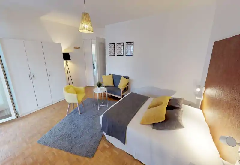 Spacious, well-staged bedroom with a large double bed, warm headboard, cozy textiles and yellow accent seating. Natural and artificial light create a bright, welcoming atmosphere ideal for rental photos.