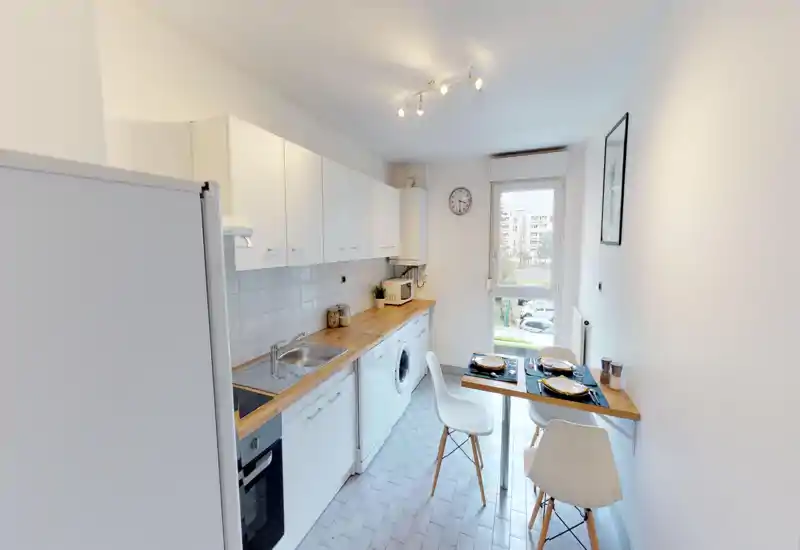 Bright, modern galley kitchen with white cabinets, wooden countertop, integrated appliances and a small dining nook by the window.