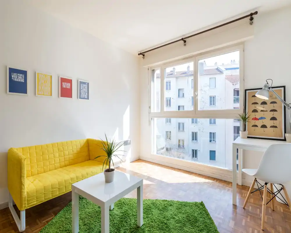 Salon moderne et lumineux avec un canapé jaune vif, tapis vert et grande fenêtre apportant une belle lumière naturelle — espace accueillant pour se détendre ou travailler.