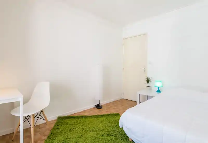 Chambre lumineuse et minimaliste avec lit blanc bien fait, tapis vert et petite table de chevet avec lampe — idéale pour des annonces locatives modernes et chaleureuses.