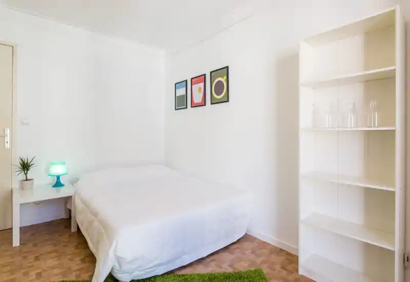 Chambre lumineuse et minimaliste avec un lit double bien fait, une table de chevet avec lampe et plante, trois cadres au mur et une étagère ouverte — idéale pour une annonce de location moderne.