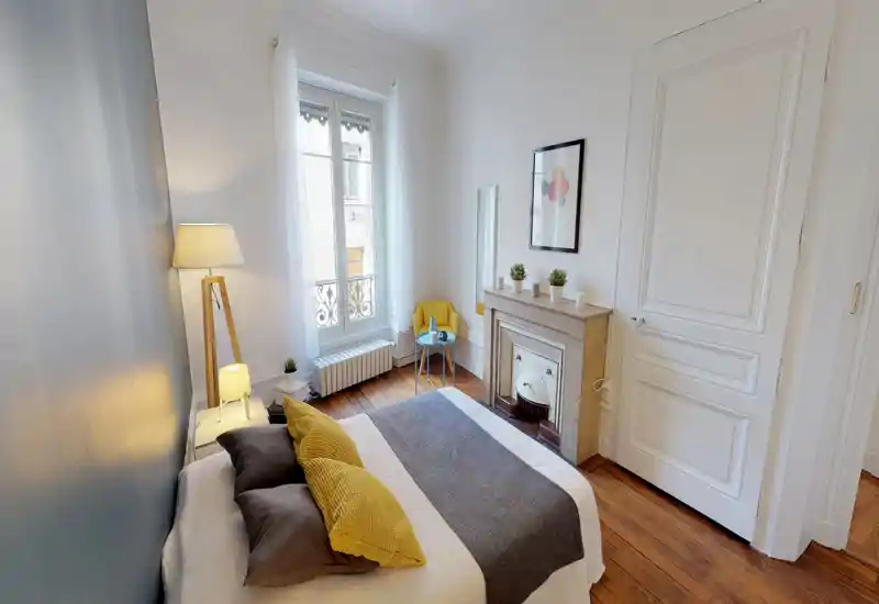 Camera da letto luminosa e curata con un letto matrimoniale ben rifatto, pavimenti in legno, camino decorativo e una grande finestra che offre luce naturale.