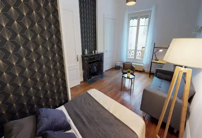 Bright, stylish bedroom with a comfy double bed in the foreground, decorative dark patterned wallpaper, wooden floors and a small seating area with sofa and coffee table by the window.