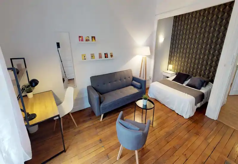 Stylish studio bedroom corner with a comfortable double bed against patterned wallpaper, a grey sofa, small seating table and a work desk — bright, modern and well-styled.