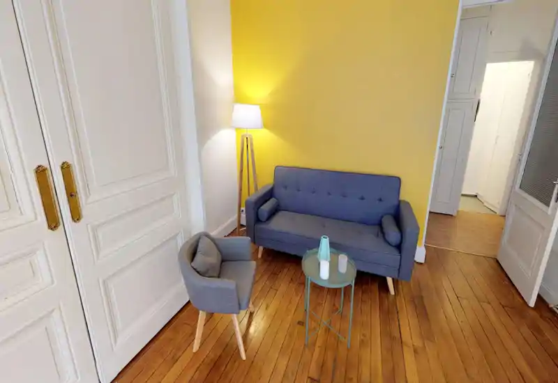 Soggiorno accogliente con divano blu, poltrona abbinata e un piccolo tavolino rotondo davanti a una parete gialla. Spazio luminoso e curato, ideale per rilassarsi o leggere.