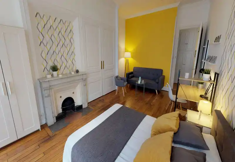 Camera da letto elegante e luminosa con un grande letto in primo piano, parete d'accento gialla, biancheria accogliente e un'area salotto con divano e poltrona — perfetta per annunci.