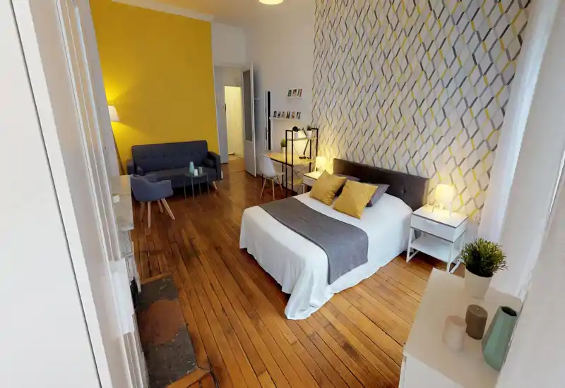 Camera da letto ampia e luminosa con letto matrimoniale dalla biancheria bianca, cuscini decorativi e plaid grigio. Pavimenti in legno, carta da parati geometrica, comodino con lampada e zona con divano e poltrona.