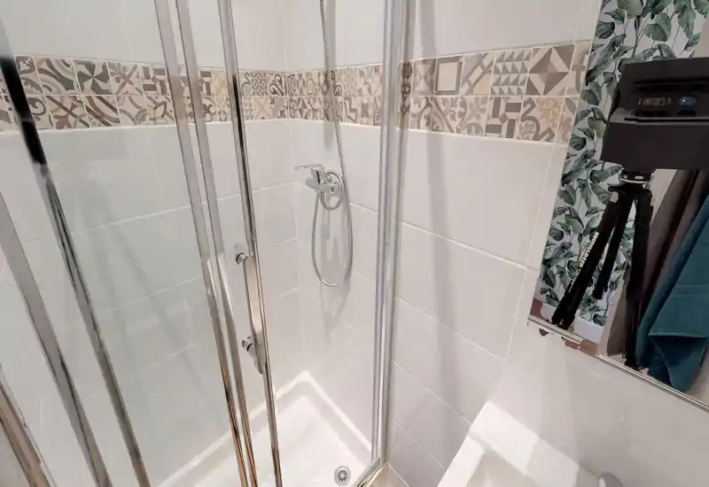 Salle de bains moderne et compacte avec cabine de douche d'angle, carrelage blanc et frise décorative; robinetterie chromée et finition lumineuse et soignée.