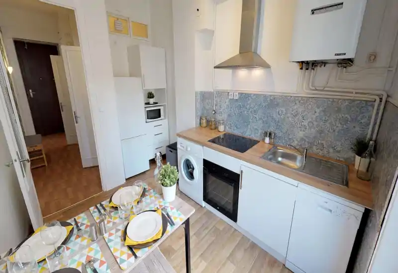Cuisine compacte et bien équipée avec plan de travail en bois, plaque à induction, four encastré, machine à laver et lave-vaisselle. Carrelage décoratif, plantes en pot et petite table à manger rendent l'espace chaleureux et pratique pour les locations courte durée.