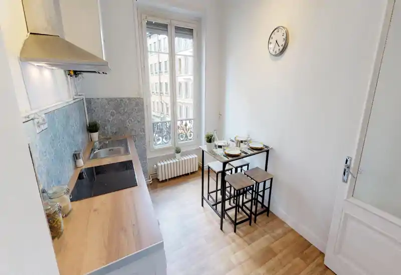 Cuisine compacte et lumineuse avec plan de travail en bois, plaque à induction, évier inox et petite table bar pour quatre près de la fenêtre; crédence à motifs et plantes apportent du charme.