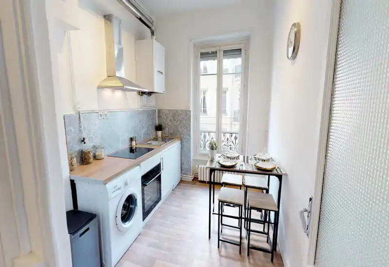 Compact, modern kitchen with integrated oven, washing machine and a small dining table by a bright window — ideal for a city flat. Decorative tile backsplash and wood-effect worktop create a warm, contemporary feel.