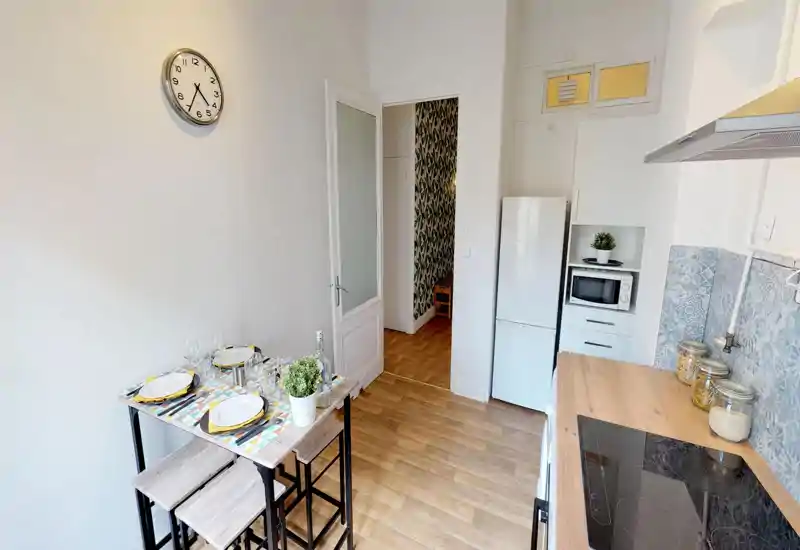 Cuisine compacte et lumineuse avec plan de travail en bois, plaque à induction, réfrigérateur et micro-ondes; un petit coin repas dressé apporte une touche chaleureuse.