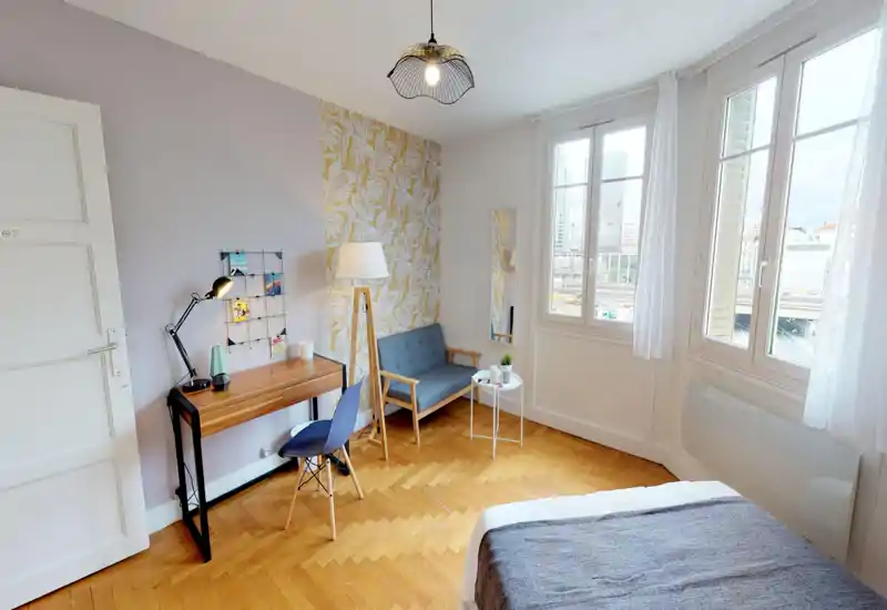 Bright, styled bedroom corner with a visible bed in the foreground, a wooden desk and chair, a small sofa with a floor lamp and large windows providing city views.