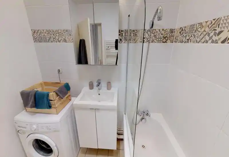 Clean, modern bathroom with a bathtub and showerhead, a white vanity sink, mirror cabinet and a washing machine. Decorative patterned tile band adds character and the space is well lit.