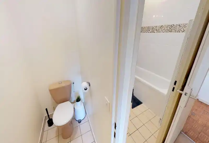 Compact, well-lit bathroom featuring a toilet and a separate bathtub with tiled walls and patterned trim.