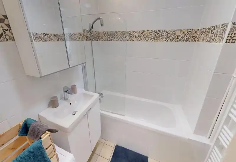 Bright, modern bathroom with a bathtub and shower screen, white tiles with decorative border, a sink cabinet and mirrored medicine cabinet.