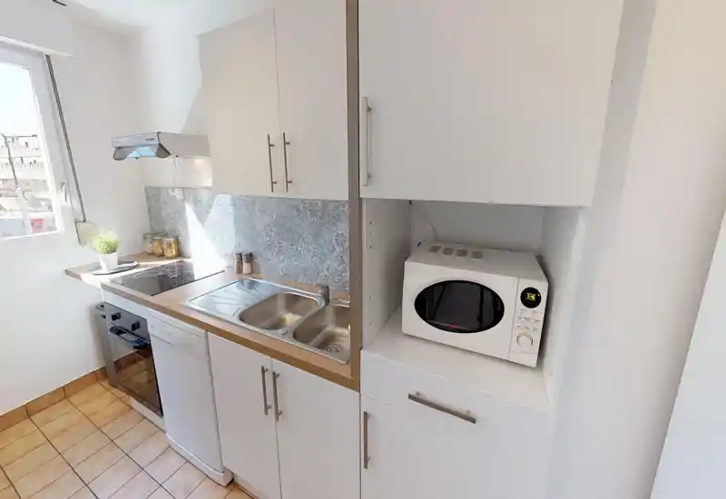 Bright compact kitchen with white cabinets, electric stove, dishwasher and built-in microwave — styled with a patterned tiled backsplash and a small potted plant.
