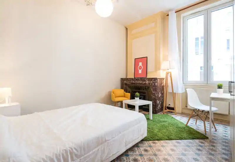 Bright, stylish bedroom with a large bed, decorative marble fireplace, cozy green rug and a small workspace by the window — ideal for a comfortable stay.
