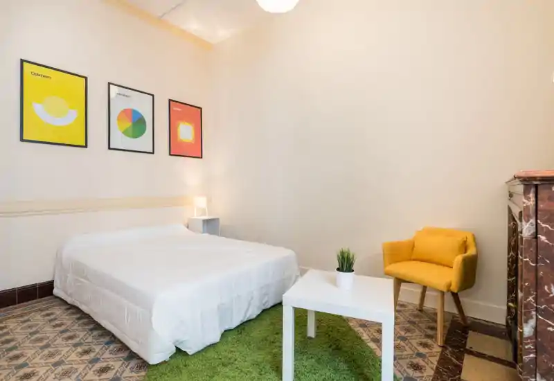 Bright, minimalist bedroom with a white double bed, colorful framed prints, a green rug and a mustard accent chair — cozy and well-lit.