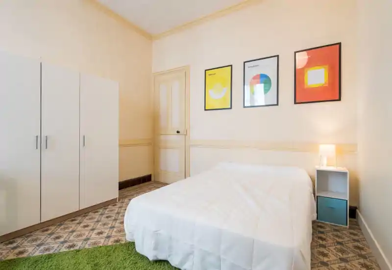 Bright, tidy bedroom with a double bed, simple bedside table and lamp, a large white wardrobe and colorful framed prints on the wall — ideal for a clean, minimalist rental listing.