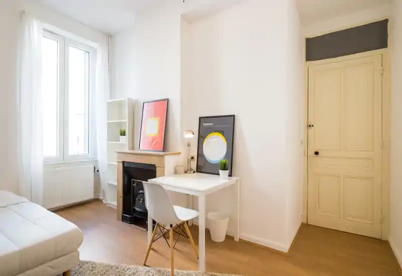 Bright, minimal bedroom with a white bed, a small desk and decorative framed prints leaning on the mantel. Large window with sheer curtains fills the room with natural light; clean wooden floor and simple modern furnishings create a calm, airy atmosphere.