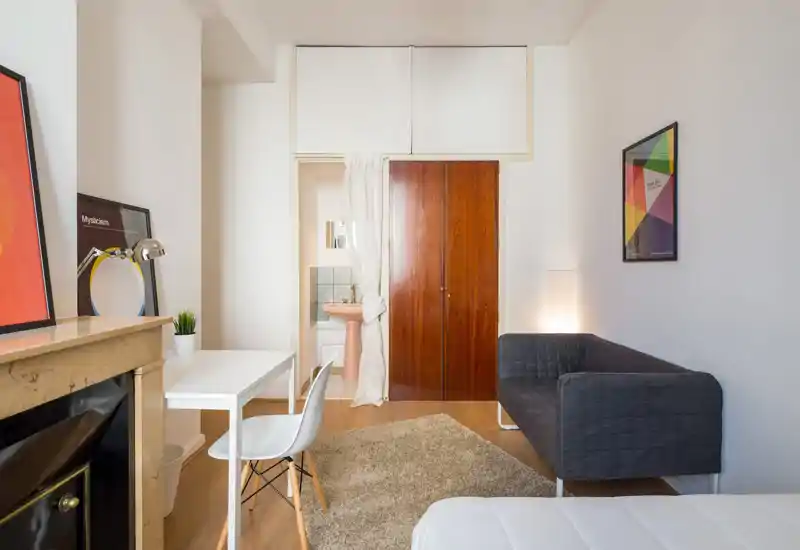 Bright, neatly styled bedroom corner with a visible white bed, a small desk and chair, a dark grey sofa and a wardrobe; warm wooden floor and soft area rug create a welcoming atmosphere.
