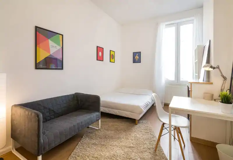 Bright, minimal bedroom with a double bed, a small sofa, desk and natural light from a large window — ideal for a cozy single or couple.