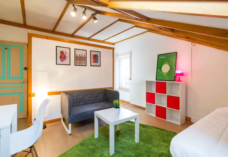 Bright, cozy living area with a compact gray sofa, white coffee table on a green rug and decorative shelving with red storage bins, staged for short-term rentals.