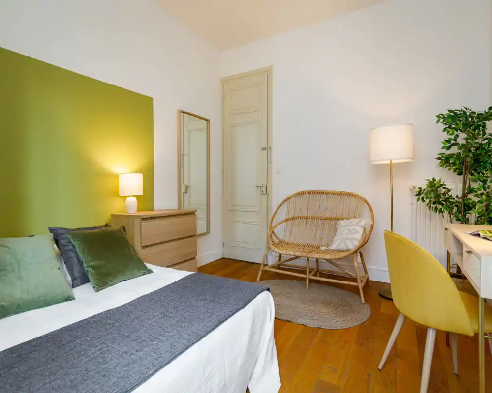 Bright, styled bedroom with a comfortable double bed, green accent wall, wooden flooring and cozy rattan seating — ideal for a relaxing stay.