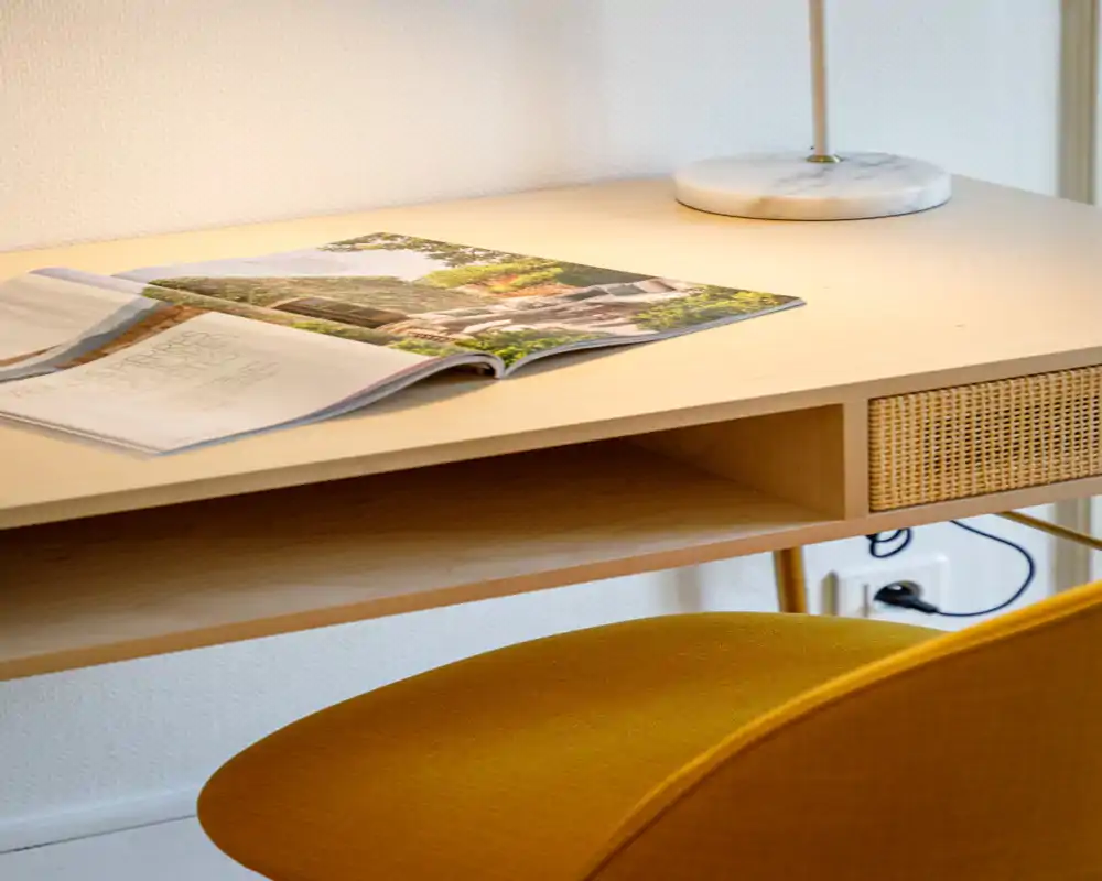 A bright, tidy sitting/working corner with a mustard upholstered chair, wooden desk, lamp and an open magazine — a cozy, well-lit nook ideal for reading or light work.