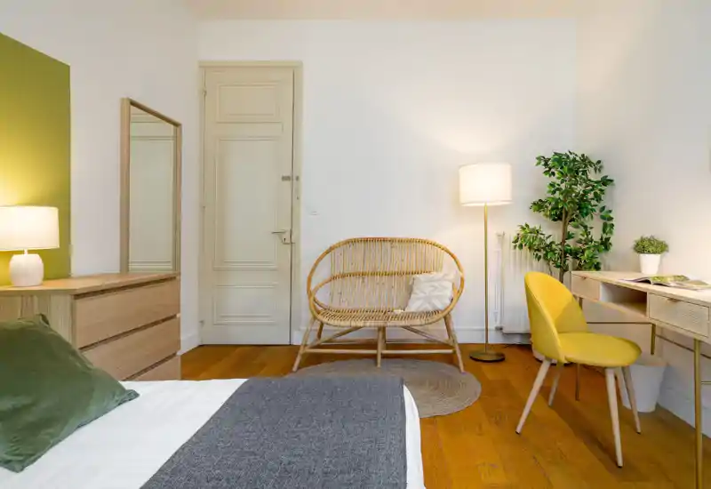 Bright, modern bedroom corner featuring a neatly made bed in the foreground, a wooden dresser with lamp, a rattan loveseat, and a small desk with a yellow chair — warm, inviting styling and natural textures.