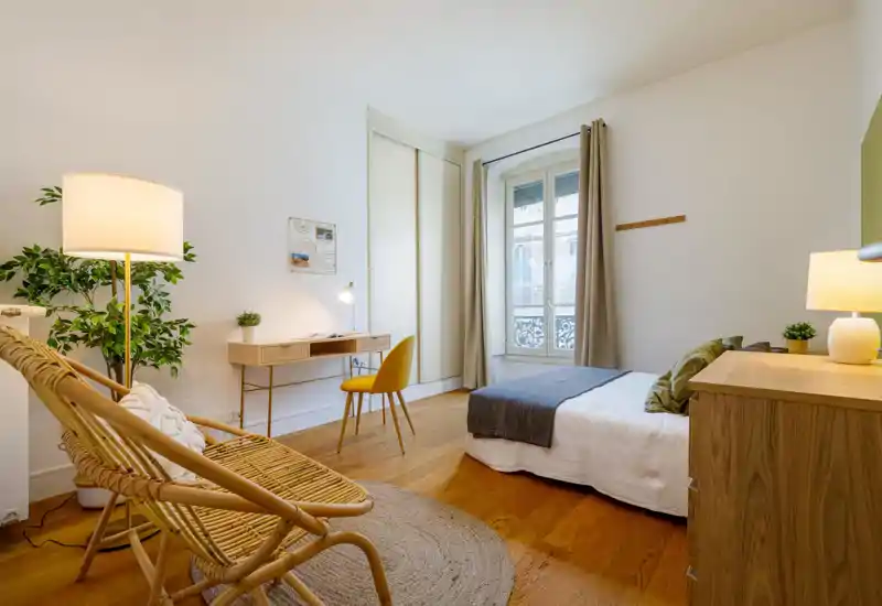 Sunny, modern bedroom with a comfortable single bed, wooden floors and a cozy reading corner with rattan chairs and a standing lamp. A small desk by the window and neutral textiles give the room a calm, well-styled look perfect for short-term rentals.