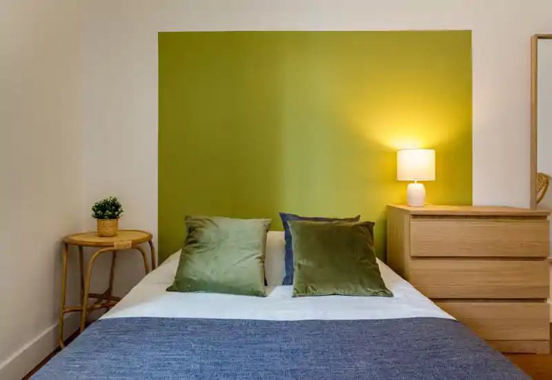 Bright, stylish bedroom with a central double bed, green accent wall and soft pillows; warm lamp light and wooden furniture create a cozy, modern atmosphere.