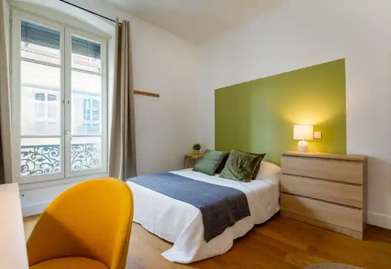 Bright, neatly styled bedroom with a double bed, green accent wall and wooden chest of drawers. Natural light from a tall window and a bedside lamp create a warm, inviting atmosphere.