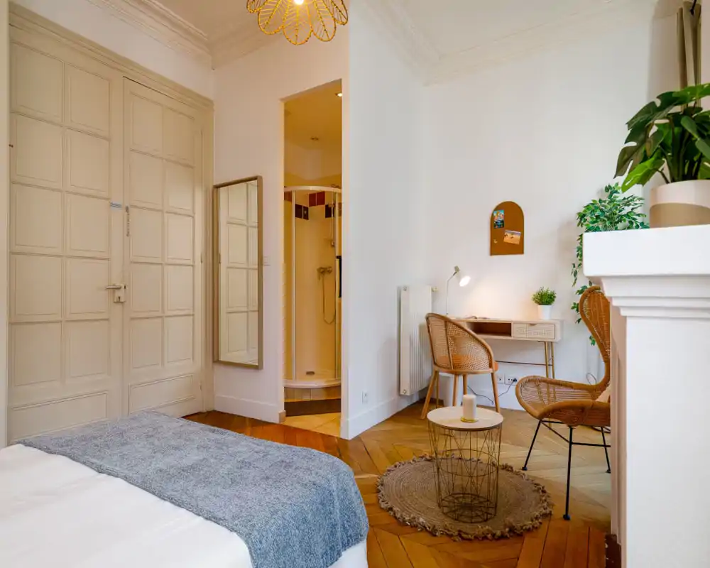 Bright, stylish bedroom corner with a visible bed in the foreground, wooden herringbone floor, wicker seating and a small desk—ideal for a cozy studio stay.