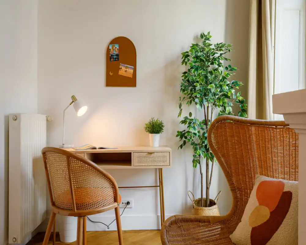 A bright, cozy living area with a rattan chair and a small desk — ideal as a work-from-home corner. Natural light, warm textiles and green plants create a welcoming atmosphere.