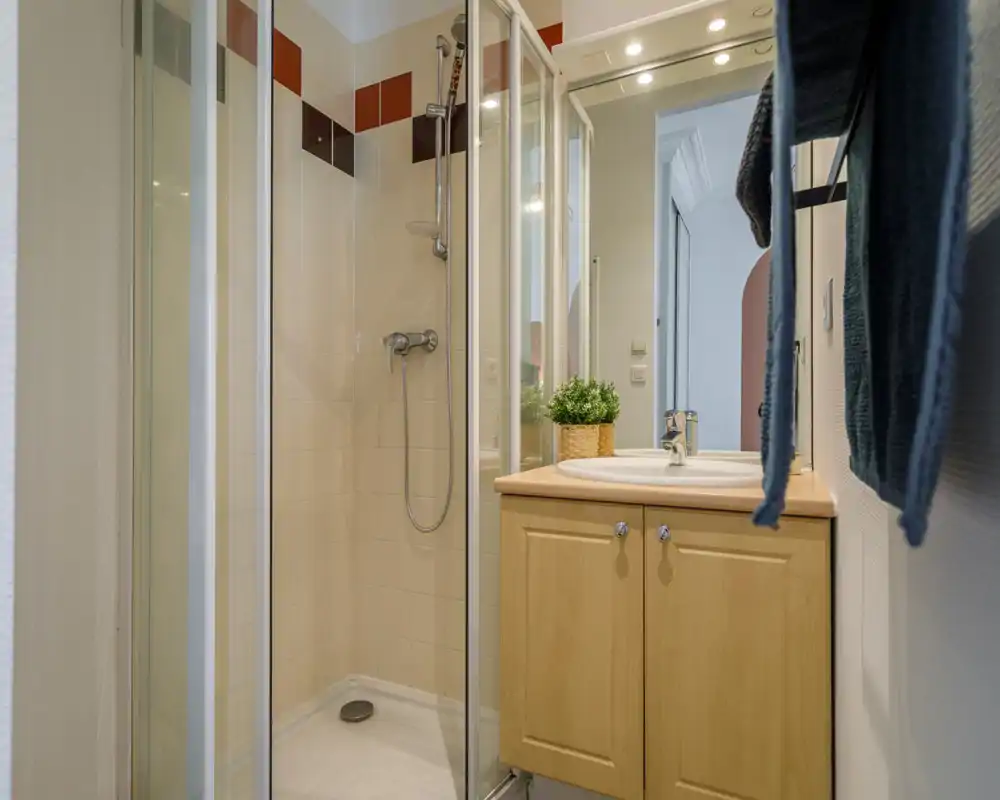 Compact, well-lit bathroom with a corner shower, sink with vanity cabinet and a mirror. Clean tiles and simple, functional fittings make it practical for guests.
