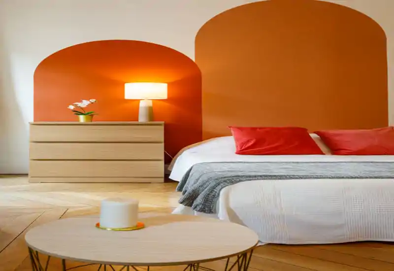 Bright, stylish bedroom with a white bed dressed with red cushions, a gray throw and a wooden chest with a table lamp; warm orange arch-painted wall adds character.