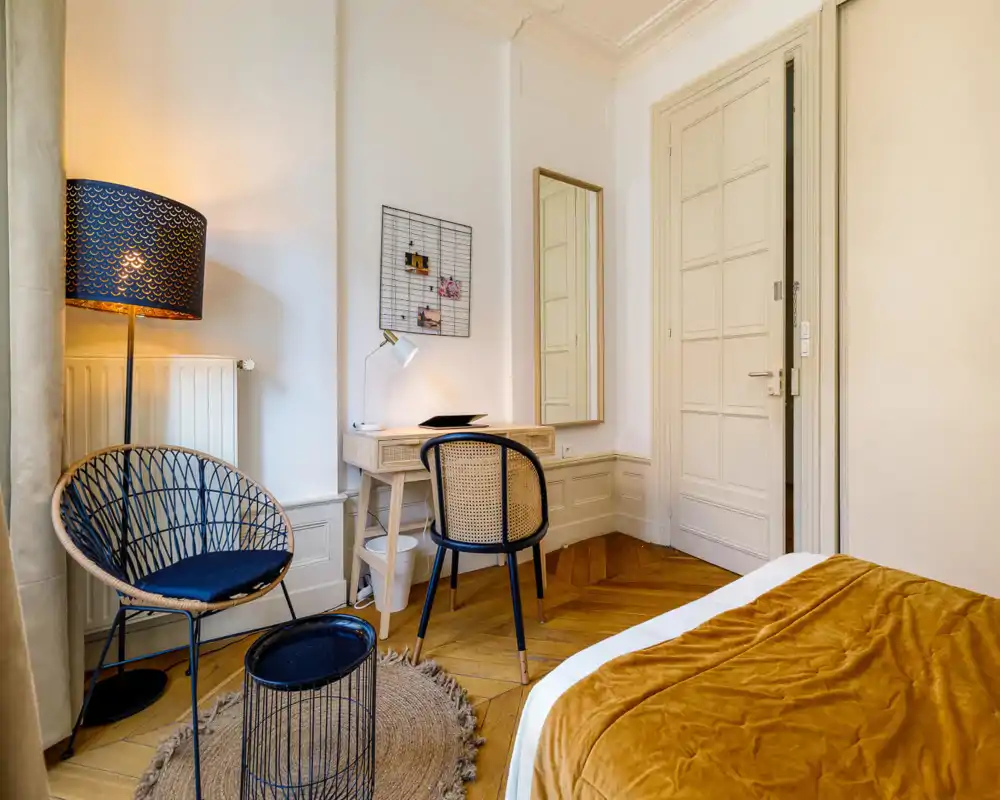 Warm, stylish bedroom corner with a partial view of the bed covered in a mustard throw, a wooden desk with chair, woven lounge chair and soft ambient lighting — ideal for a cozy work-and-relax space.