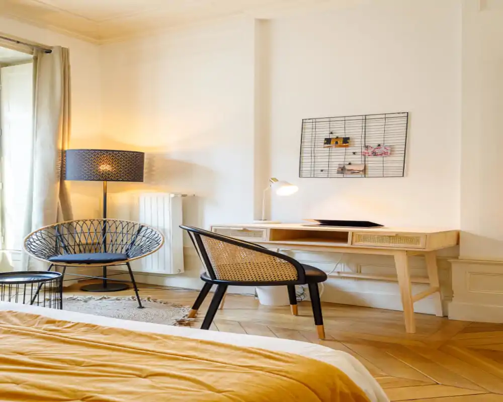 Sunny, stylish bedroom corner with a visible bed in warm ochre bedding, a wooden desk and rattan-accent chair — bright natural light and cozy lamps create an inviting workspace and reading nook.