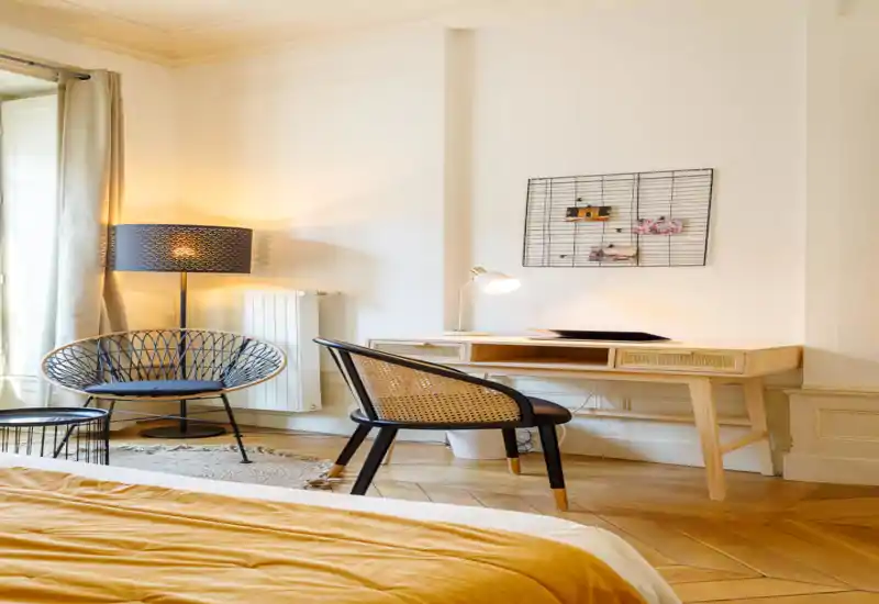 Sunny, stylish bedroom corner with a visible bed in warm ochre bedding, a wooden desk and rattan-accent chair — bright natural light and cozy lamps create an inviting workspace and reading nook.